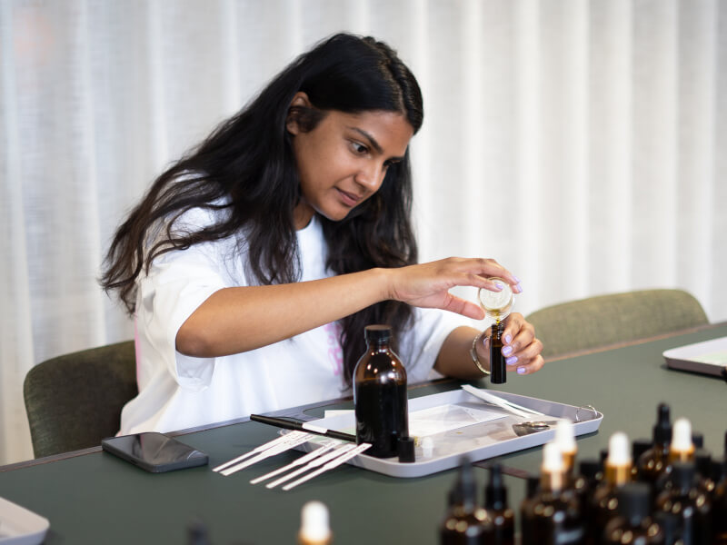 A woman is sitting at a table, pouring essential oils from a beaker into an amber glass bottle